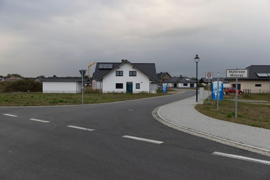 Eine Straße führt in einen Ort mit mehreren neuen Häusern. Rechts neben der Straße steht ein Schild mit der Aufschrift Mühlrose.