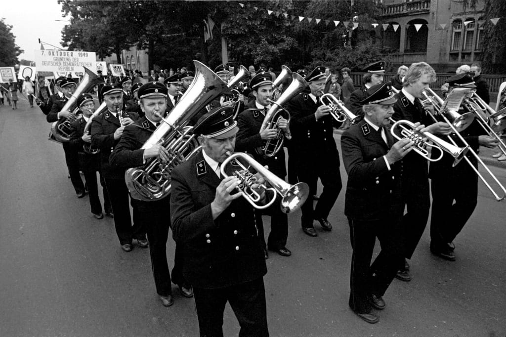 Eine Marschkapelle aus uniformierten Männern mit Blasinstrumenten läuft eine Straße entlang und spielt Musik.